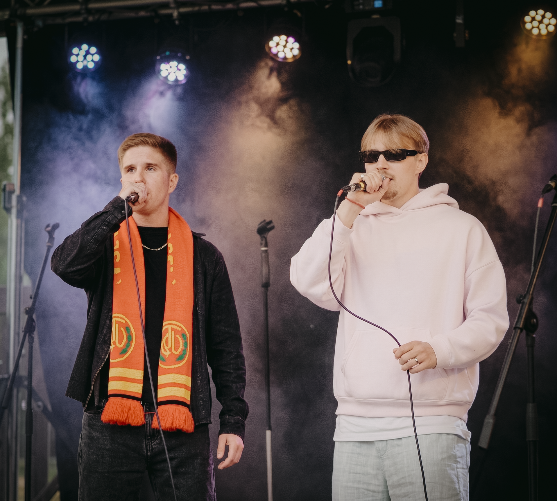 Kaksi miestä esiintyy lavalla, toinen laulaa mikrofoniin, toinen soittaa, taustalla valot ja savua.
